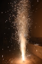 An intense burst of fireworks erupts from the ground, emitting a cascade of bright sparks and trails of light against a dark background. The fiery spectacle illuminates the immediate surroundings, creating a visually captivating display.