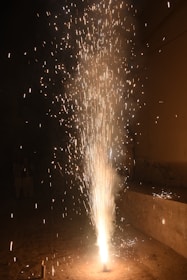 An intense burst of fireworks erupts from the ground, emitting a cascade of bright sparks and trails of light against a dark background. The fiery spectacle illuminates the immediate surroundings, creating a visually captivating display.