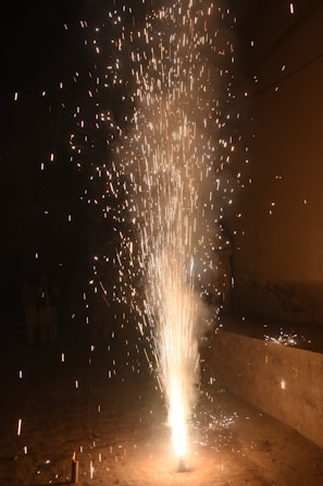 An intense burst of fireworks erupts from the ground, emitting a cascade of bright sparks and trails of light against a dark background. The fiery spectacle illuminates the immediate surroundings, creating a visually captivating display.