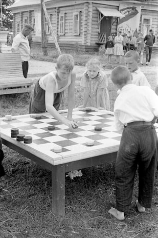 Children are gathered around an outdoor table playing checkers in front of a wooden building. The kids seem to be focused and involved in the game. In the background, a few adults and children are casually standing and observing the scene. The setting appears to be in a rural area, with a simple wooden structure, possibly a house, and some grass visible around.