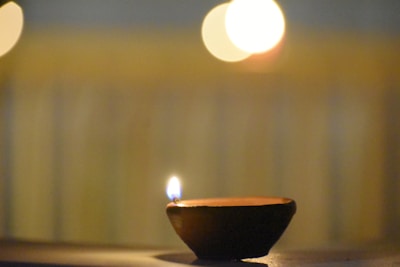 A small clay lamp with a single flame is placed on a flat surface. The background features soft, blurred lights and vertical lines, giving the impression of a warm, serene atmosphere.