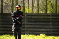 A person wearing tactical gear, including a helmet and mask, is holding a paintball marker while standing on a grassy field in front of a wooden barrier. Sunlight filters through the trees in the background, casting dappled shadows.