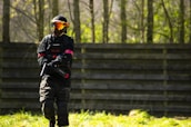 A person wearing tactical gear, including a helmet and mask, is holding a paintball marker while standing on a grassy field in front of a wooden barrier. Sunlight filters through the trees in the background, casting dappled shadows.