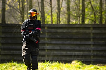 A person wearing tactical gear, including a helmet and mask, is holding a paintball marker while standing on a grassy field in front of a wooden barrier. Sunlight filters through the trees in the background, casting dappled shadows.
