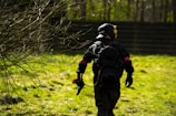 A person wearing tactical gear and a helmet is walking through a grassy area with scattered bare branches. They carry a paintball gun and are dressed in dark clothing with a red armband. The background includes a fence and several trees.