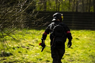 A person wearing tactical gear and a helmet is walking through a grassy area with scattered bare branches. They carry a paintball gun and are dressed in dark clothing with a red armband. The background includes a fence and several trees.