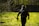 A person wearing tactical gear and a helmet is walking through a grassy area with scattered bare branches. They carry a paintball gun and are dressed in dark clothing with a red armband. The background includes a fence and several trees.