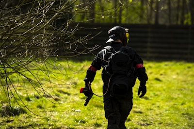 A person wearing tactical gear and a helmet is walking through a grassy area with scattered bare branches. They carry a paintball gun and are dressed in dark clothing with a red armband. The background includes a fence and several trees.