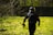 A person wearing tactical gear and a helmet is walking through a grassy area with scattered bare branches. They carry a paintball gun and are dressed in dark clothing with a red armband. The background includes a fence and several trees.