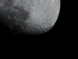A close-up of the Moon's surface, revealing numerous craters and rough textures. The lunar surface is partially illuminated against a pitch-black sky, highlighting its rugged and cratered appearance.