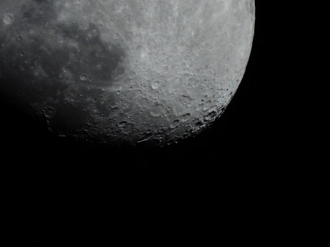 A close-up of the Moon's surface, revealing numerous craters and rough textures. The lunar surface is partially illuminated against a pitch-black sky, highlighting its rugged and cratered appearance.