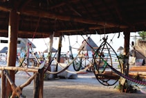 A tropical beach setting with a thatched-roofed hut featuring several colorful hammocks and a hanging chair. In the background, there are people walking, bicycles, and beach huts with thatched roofs. The area is well-lit with natural light, suggesting a lively, sunny day.