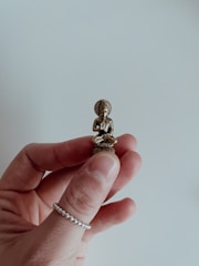 A small Buddha sculpture is delicately held between two fingers. The sculpture is made of a brass-like material with intricate details visible on the figure's robe and facial features. The background is a soft, out-of-focus light gray, providing a neutral contrast to the metallic tones of the sculpture.