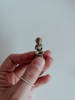 A small Buddha sculpture is delicately held between two fingers. The sculpture is made of a brass-like material with intricate details visible on the figure's robe and facial features. The background is a soft, out-of-focus light gray, providing a neutral contrast to the metallic tones of the sculpture.