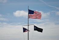 Three flags are mounted on a pole against a backdrop of a blue sky with scattered clouds. The largest flag is the United States flag, prominently displayed at the top. Below it, there is a state flag, partially visible, and another dark flag with the words 'Marina at Caesar Creek' printed on it.