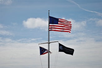 Three flags are mounted on a pole against a backdrop of a blue sky with scattered clouds. The largest flag is the United States flag, prominently displayed at the top. Below it, there is a state flag, partially visible, and another dark flag with the words 'Marina at Caesar Creek' printed on it.