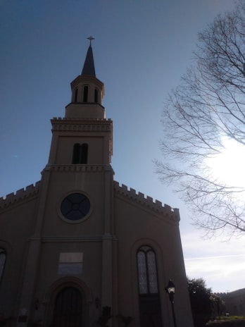 A church building with a tall steeple topped by a cross is situated under a clear blue sky. There is a bare tree with branches extending to the right side, with the sun shining brightly behind it, casting long shadows.