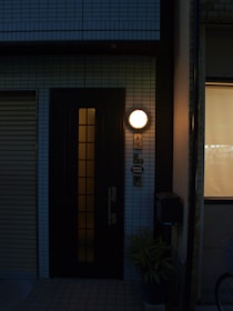 A dimly lit entryway featuring a dark door with glass panels, illuminated by a round exterior light above. The surrounding wall is tiled, and there is a small potted plant at the base. An adjacent window with a blind partially drawn is visible, along with some signage indicating an address.