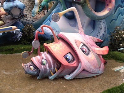 A colorful and whimsical sculpture of a fish with exaggerated features and large eyes, resting on a pathway. The fish is adorned with various artistic patterns and seems to incorporate other smaller fish within its design. Surrounding the sculpture are elements that resemble vegetation and other abstract structures.