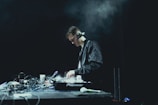 A person wearing headphones is focused on operating a DJ mixer, surrounded by various cables and electronic equipment on a dimly lit stage. The background is dark, enhancing the concentration and seriousness of the environment.