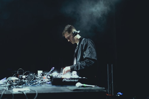 A person wearing headphones is focused on operating a DJ mixer, surrounded by various cables and electronic equipment on a dimly lit stage. The background is dark, enhancing the concentration and seriousness of the environment.