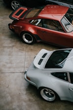 Three classic sports cars are parked closely together in an indoor setting. The cars feature sleek designs with curved bodies and prominent wheel arches. The floor is a polished concrete.