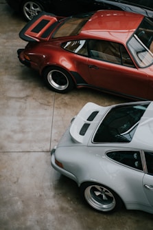 Three classic sports cars are parked closely together in an indoor setting. The cars feature sleek designs with curved bodies and prominent wheel arches. The floor is a polished concrete.