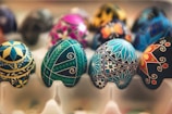 Intricately decorated eggs featuring detailed geometric patterns and vibrant colors are arranged in a tray. The designs include stars, floral motifs, and intricate line work with a mix of blues, greens, yellows, blacks, and other bright hues.