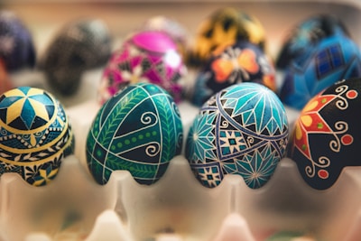 Intricately decorated eggs featuring detailed geometric patterns and vibrant colors are arranged in a tray. The designs include stars, floral motifs, and intricate line work with a mix of blues, greens, yellows, blacks, and other bright hues.