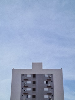 A simple high-rise building is centered against a vast, clear sky. The building exhibits a minimalist design with a grey facade, multiple windows, and several air conditioning units attached to the exterior.