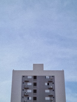 A simple high-rise building is centered against a vast, clear sky. The building exhibits a minimalist design with a grey facade, multiple windows, and several air conditioning units attached to the exterior.