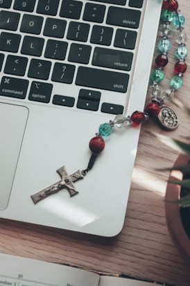 A laptop keyboard is visible with part of the trackpad in view. On the trackpad rests a rosary with colorful beads and a crucifix, along with a medallion. The wooden surface is partially visible with a book at the edge, accompanied by a plant pot in the corner.