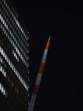 A high-rise building is visible on the left side, with its windows illuminated against a dark night sky. A tall construction crane extends diagonally, painted in alternating segments of white and orange.
