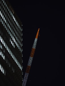 A high-rise building is visible on the left side, with its windows illuminated against a dark night sky. A tall construction crane extends diagonally, painted in alternating segments of white and orange.