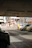The image depicts a parking lot with several cars parked. In the background, there is a street with more vehicles and various buildings, including a multi-story structure. Overhead lights are visible on the ceiling of the parking area. There is some signage visible, including a large advertisement board.