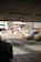 The image depicts a parking lot with several cars parked. In the background, there is a street with more vehicles and various buildings, including a multi-story structure. Overhead lights are visible on the ceiling of the parking area. There is some signage visible, including a large advertisement board.