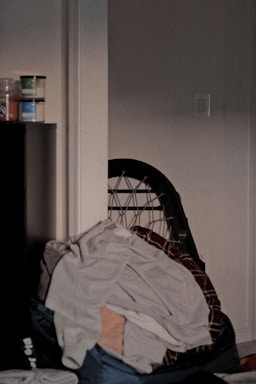 A cluttered corner of a room with a pile of clothes draped over a chair. Next to it, there are a few stacked containers or jars on a piece of furniture. The lighting is dim, giving the space a slightly disorganized and moody atmosphere.