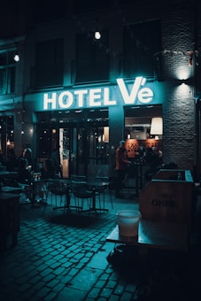 Neon-lit hotel entrance with a modern sign reading 'HOTEL V&Eacute;', outdoor seating area with tables and chairs under dim lighting. People are gathered inside and outside, with a cozy ambiance highlighted by moody lighting.