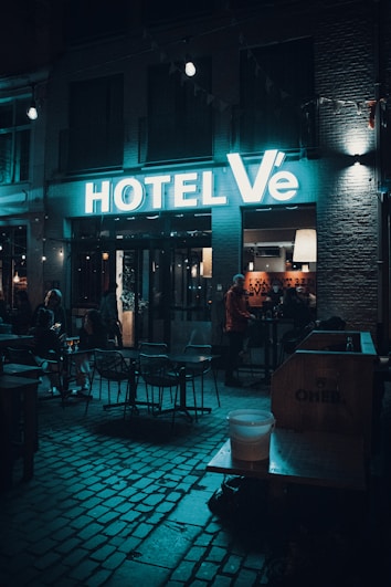 Neon-lit hotel entrance with a modern sign reading 'HOTEL V&Eacute;', outdoor seating area with tables and chairs under dim lighting. People are gathered inside and outside, with a cozy ambiance highlighted by moody lighting.