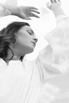 A woman with flowing dark hair is seen in a serene pose, with one arm raised above her head and eyes gently closed. She wears a loose white blouse and a delicate necklace with a small star pendant. The background appears to be a bright, overcast sky.