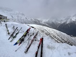 Skis and ski poles are propped up in the snow near a building at a snowy mountain slope. The background features a range of snow-covered mountains under an overcast sky.