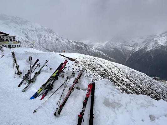 Skis and ski poles are propped up in the snow near a building at a snowy mountain slope. The background features a range of snow-covered mountains under an overcast sky.