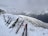 Skis and ski poles are propped up in the snow near a building at a snowy mountain slope. The background features a range of snow-covered mountains under an overcast sky.
