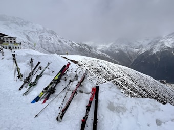 Skis and ski poles are propped up in the snow near a building at a snowy mountain slope. The background features a range of snow-covered mountains under an overcast sky.