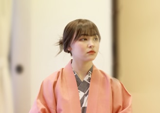 A person with a serene expression wearing a traditional Japanese kimono. The kimono is pink with subtle patterns, and the individual's hair is styled in a simple updo. The background is softly blurred, suggesting an indoor setting with warm lighting.