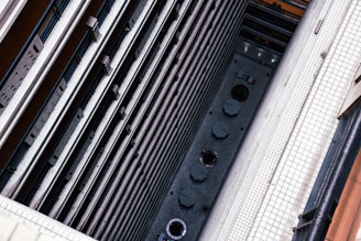 A vertical view from a high angle showing the interior of a tall building. The structure features multiple layers of balconies with railings running parallel to each other. At the ground level, tables with umbrellas and circular architectural elements are visible. The walls are covered with white tiles, and the scene is captured from a steep perspective.