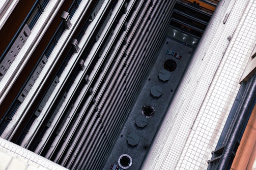 A vertical view from a high angle showing the interior of a tall building. The structure features multiple layers of balconies with railings running parallel to each other. At the ground level, tables with umbrellas and circular architectural elements are visible. The walls are covered with white tiles, and the scene is captured from a steep perspective.