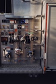 A detailed view of a fire truck control panel with various levers, knobs, and gauges. The panel features metal components and switches, some labeled in Japanese, with a prominent valve and chain in the center. The background includes a textured metal surface and part of a roller door.