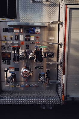 A detailed view of a fire truck control panel with various levers, knobs, and gauges. The panel features metal components and switches, some labeled in Japanese, with a prominent valve and chain in the center. The background includes a textured metal surface and part of a roller door.