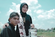A woman and a man dressed in traditional attire stand together outdoors under a bright blue sky with scattered clouds. The woman wears a hijab and patterned sash, and the man wears a traditional headpiece. They are both looking towards the camera, and the woman has her arm on the man's shoulder. In front of them, there is a stack of metal containers with floral decorations.